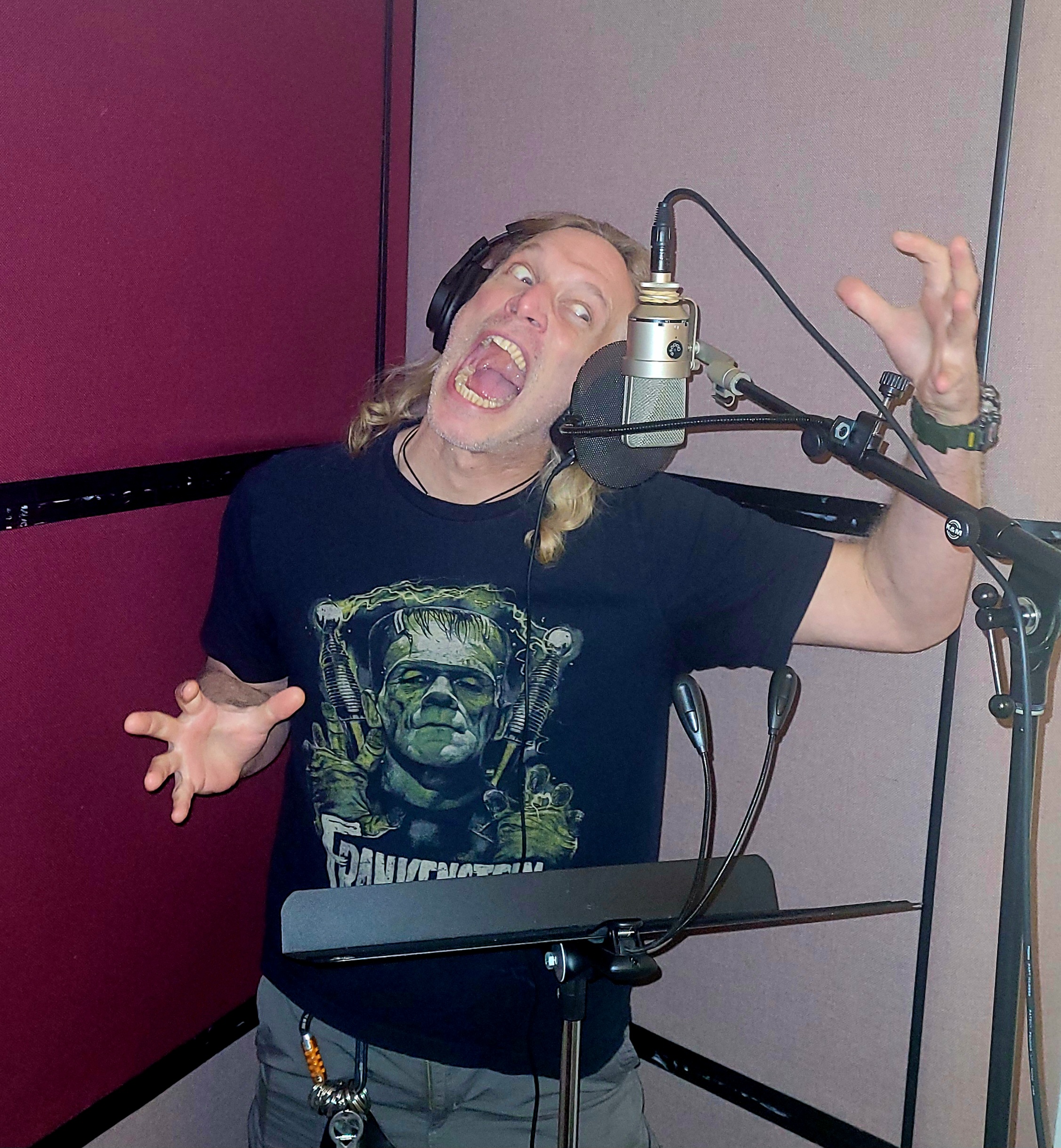 Brian Albert wearing headphones and a Frankenstein shirt, raising his hands in a monster pose beside a studio microphone.