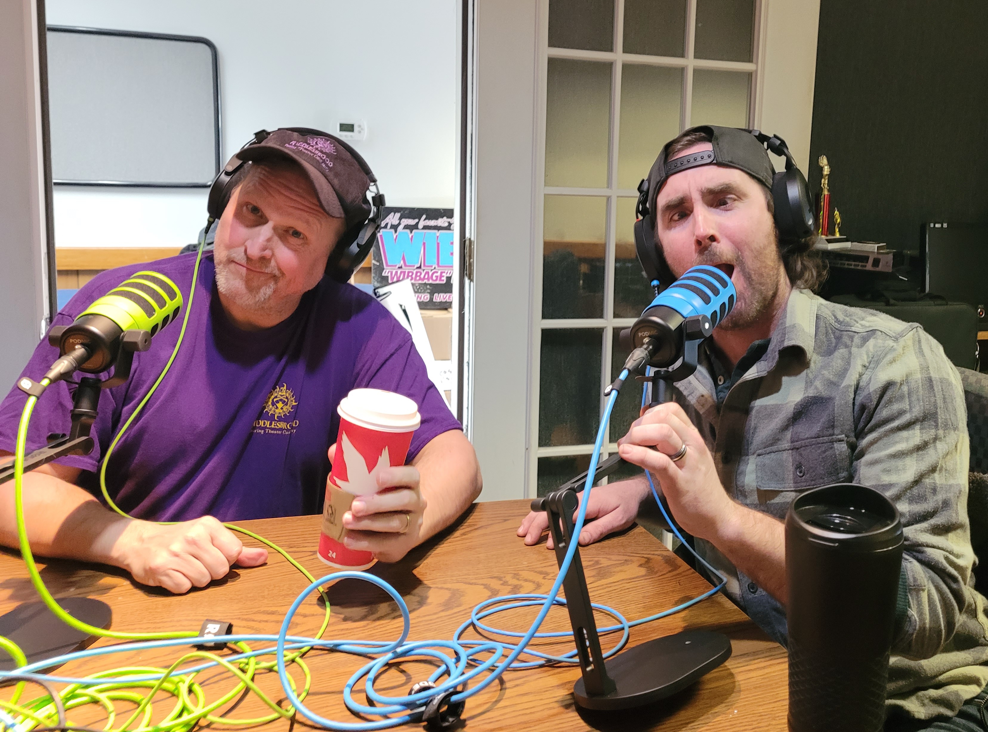 Clyde P. Riddlesbrood holding a coffee cup beside Kevin Ebner, both wearing headphones at a table with green and blue microphones.
