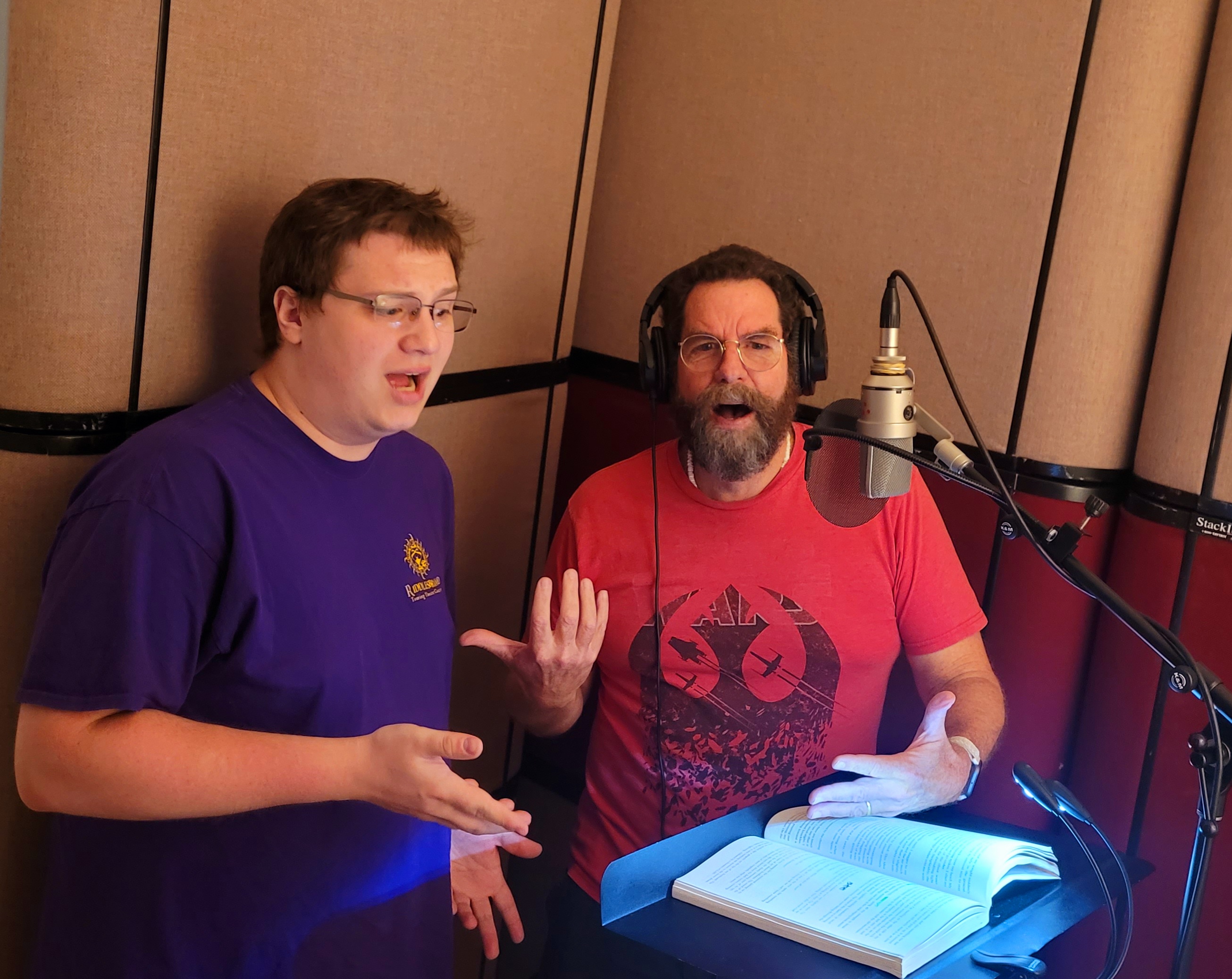 Jeff Cleve and Barry Berglund gesturing expressively beside a studio microphone and an open book during a Riddlesbrood audiobook recording session.