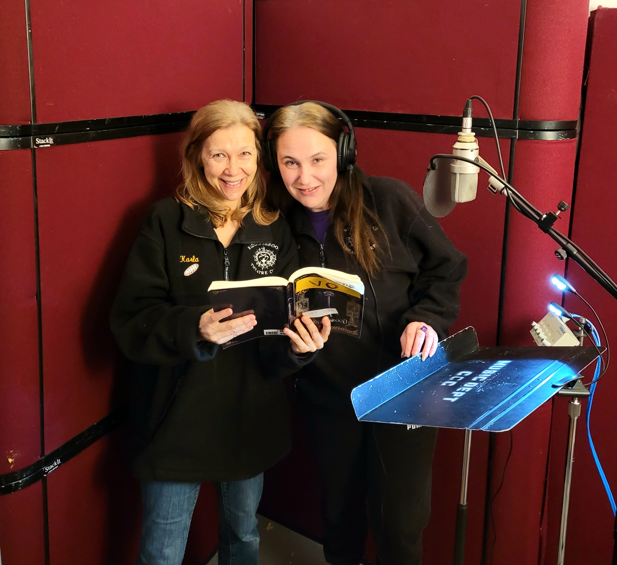 Karla Shantz and Kate Brubaker smiling beside a studio microphone with an open copy of The Greatest Brochure in the World.