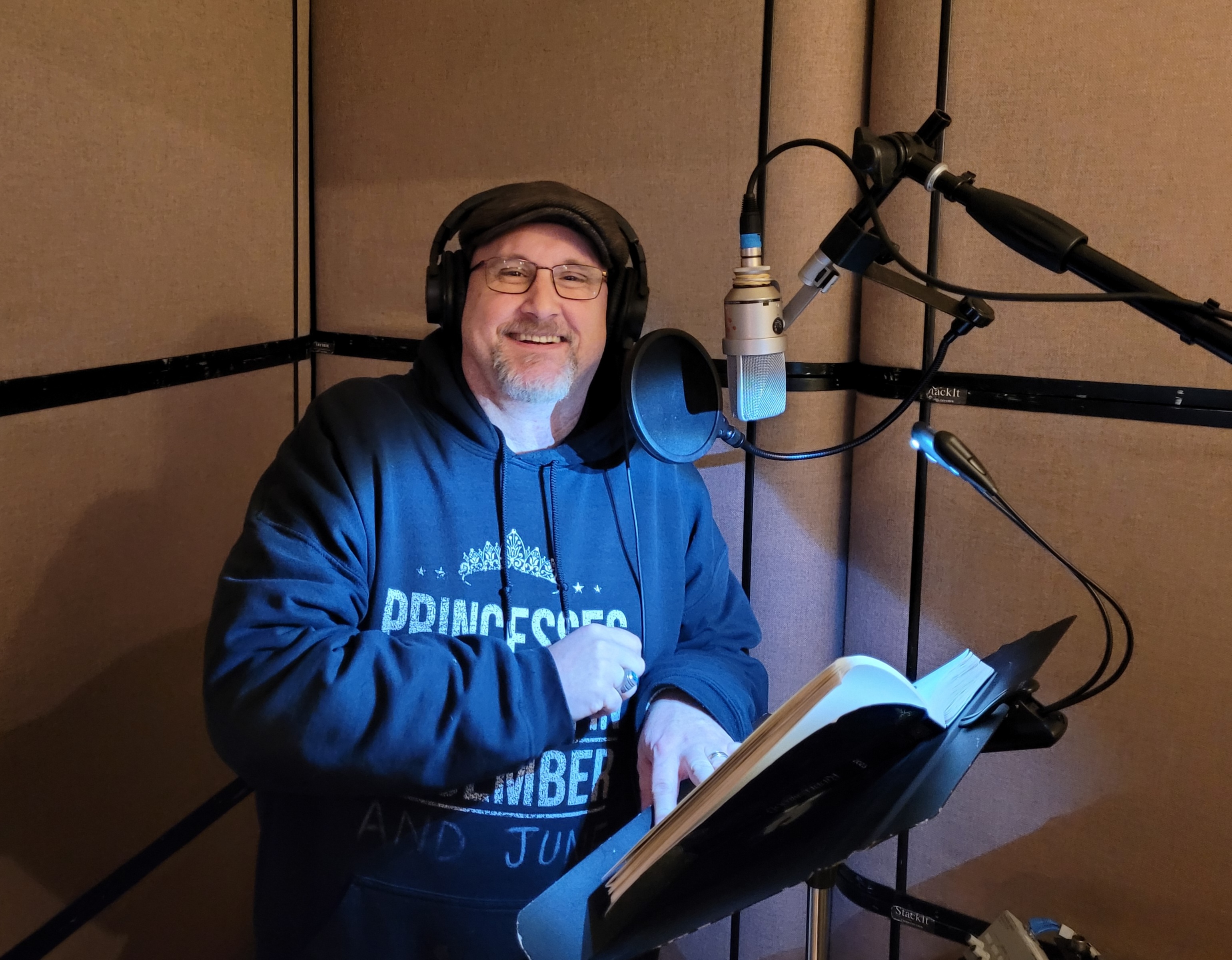 Chris Nagle smiling beside a studio microphone, wearing headphones and a flat cap, with an open book on the music stand.