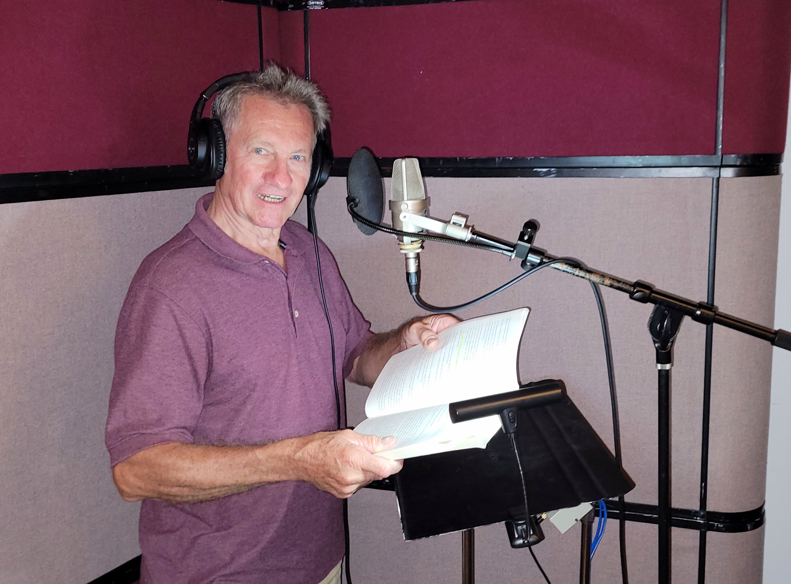 Dean “Woody” Duncan wearing headphones and holding an open book at a studio microphone during a Riddlesbrood audiobook recording session.