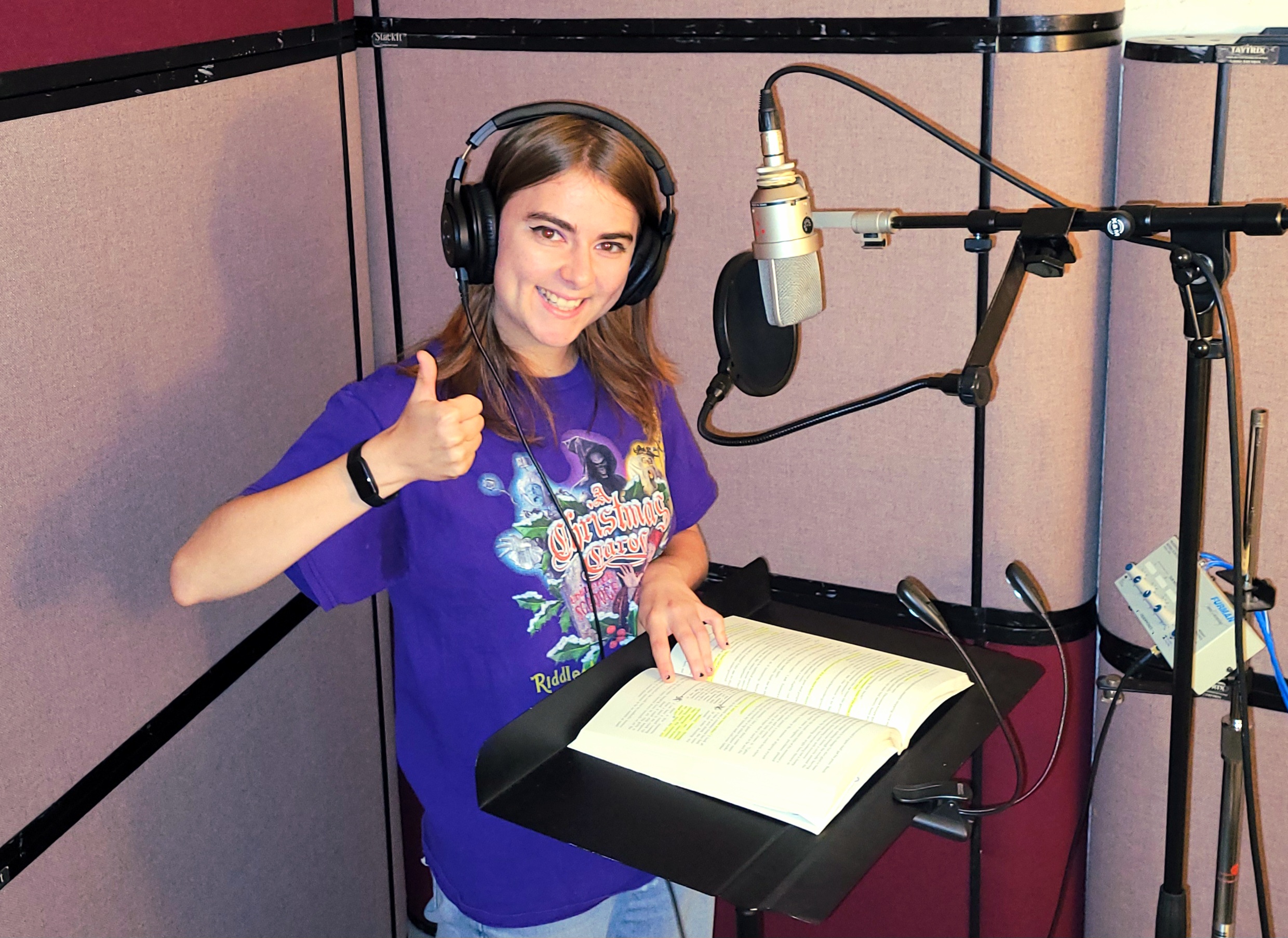 Eileen Morrison wearing headphones and giving a thumbs-up beside a studio microphone, with an open, highlighted book on the music stand.