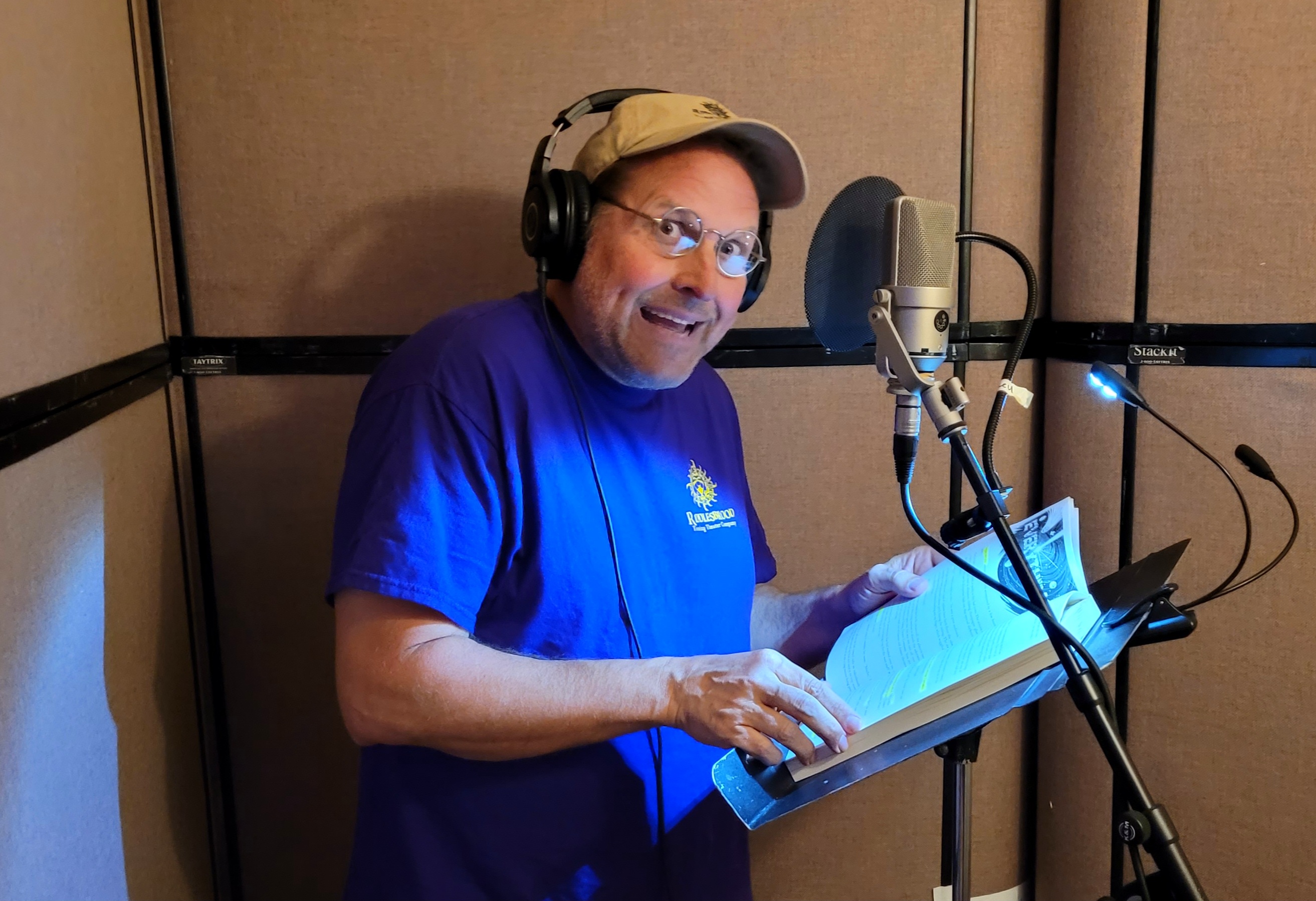 Clyde P. Riddlesbrood wearing headphones and holding an open book beside a studio microphone while recording the audiobook.